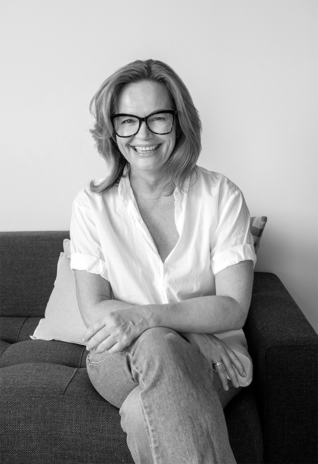 Black and white portrait of Claudia Mahler sitting on a sofa, wearing a white shirt, jeans, and glasses